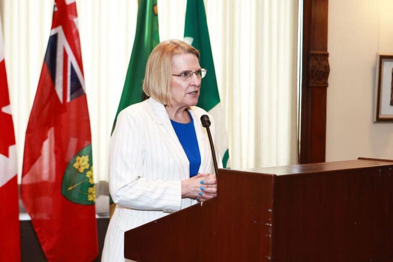 Speaker addressing a presentation at Queen's Park, Toronto, with Ontario and Canadian flags behind the podium