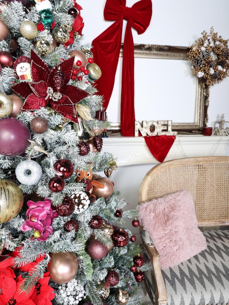 Close-up of a flocked Christmas tree decorated with burgundy poinsettias, wine and rose-gold baubles beside a red velvet bow and NOEL mantel